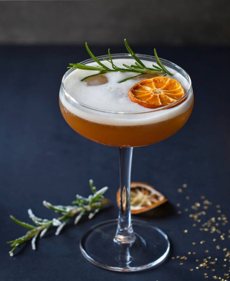 a cocktail in a stemmed glass garnished with rosemary and a slice of orange