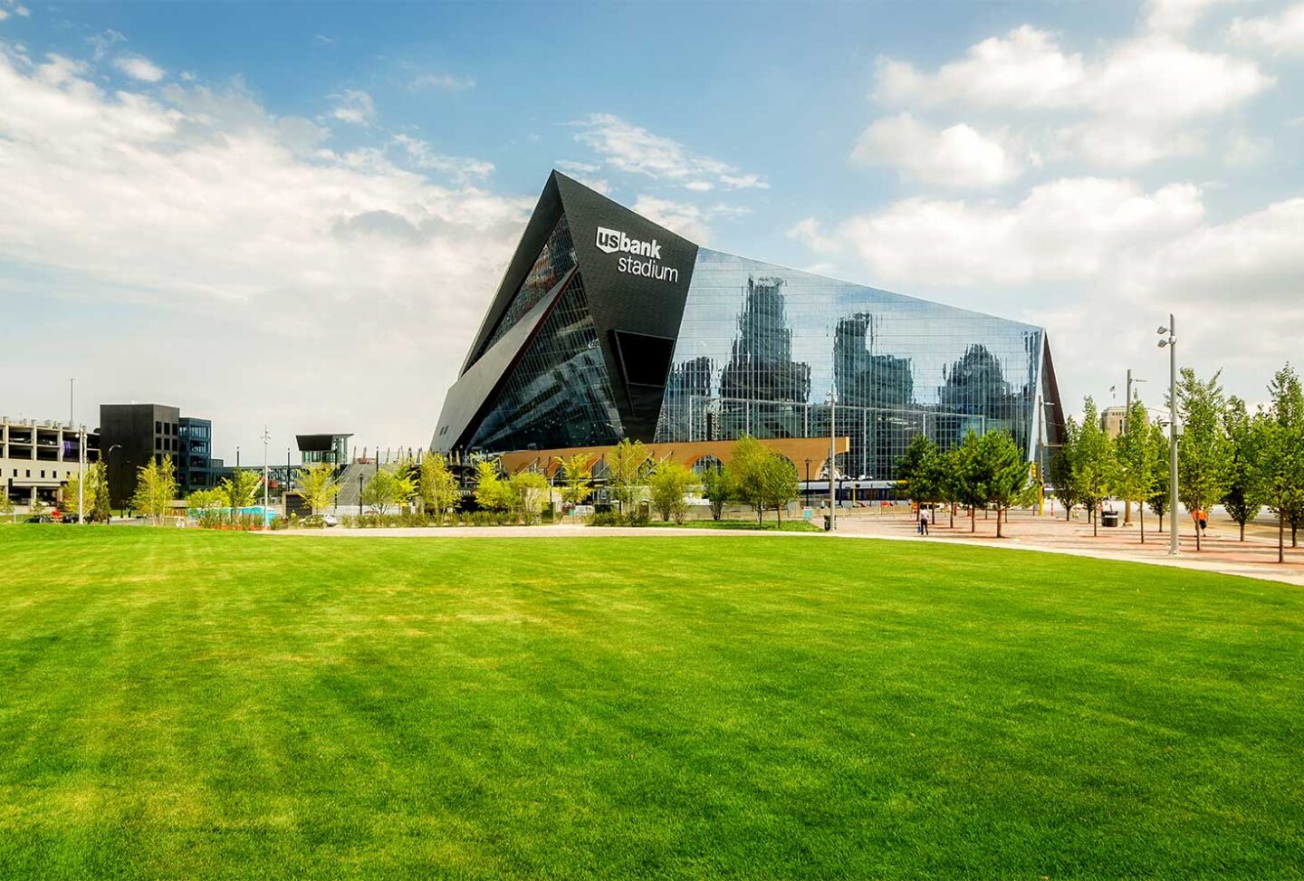 A modern glass-fronted stadium labeled U.S. Bank Stadium rises behind a large, green, open lawn with scattered trees in Minneapolis under a partly cloudy sky.