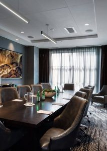A modern conference room with a long Tavola-style table, leather chairs, bottled water, notepads, and a large window with sheer curtains—ideal for meetings or private dining.