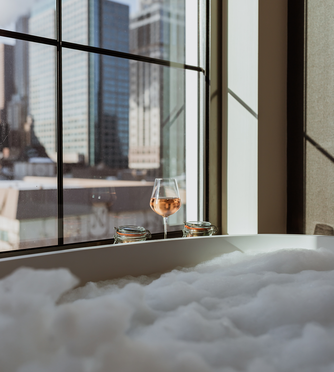 A glass of rosé sits on a windowsill next to a bubble-filled bathtub, with tall city buildings visible through the window in daylight.