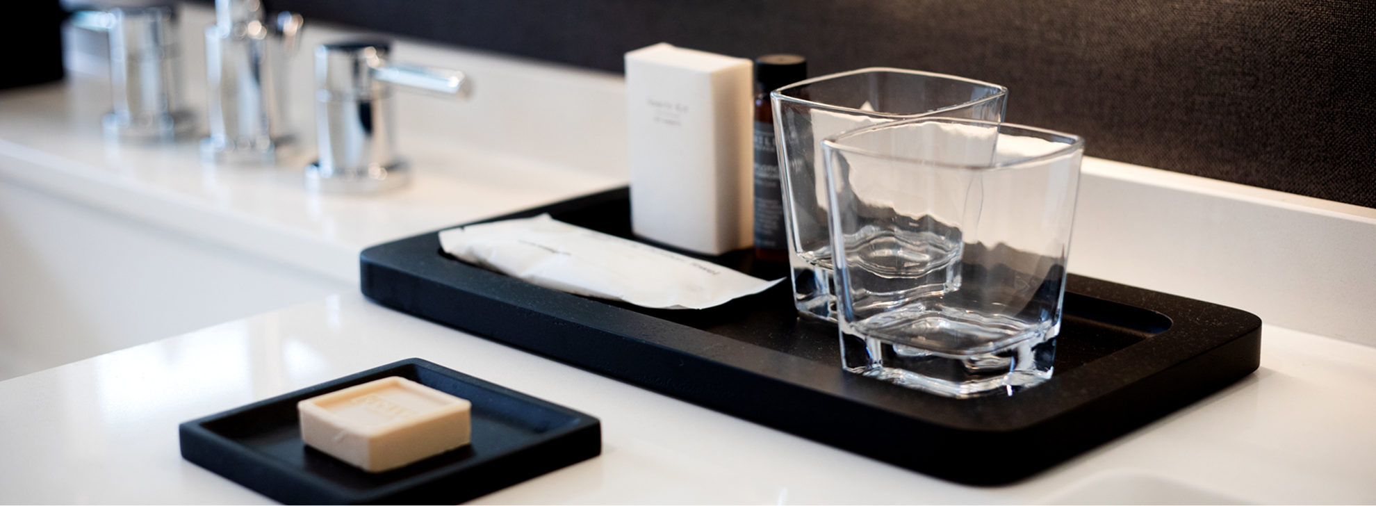 Two glass tumblers, a bar of soap, and toiletries are arranged neatly on a black tray on a white bathroom countertop.