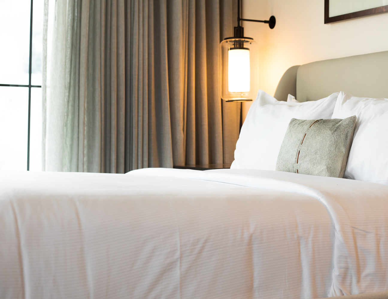 A neatly made bed with white linens and a decorative pillow, beside a lit wall lamp and floor-to-ceiling curtains.