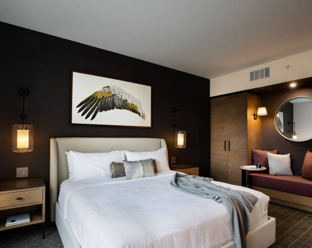 Modern hotel room with a large bed, white linens, bedside tables, a dark accent wall with a bird wing artwork, a round mirror, and a seating area with cushions.