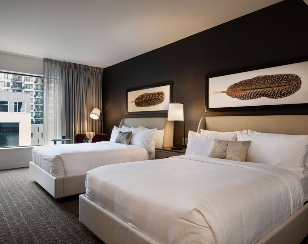A modern hotel room with two double beds, white bedding, a dark accent wall, feather artwork, a window with city view, and a floor lamp.