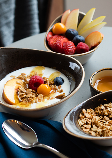 A bowl of yogurt with granola and fruit, a bowl of mixed fruit, a bowl of granola, a cup of honey, and a spoon on a table.