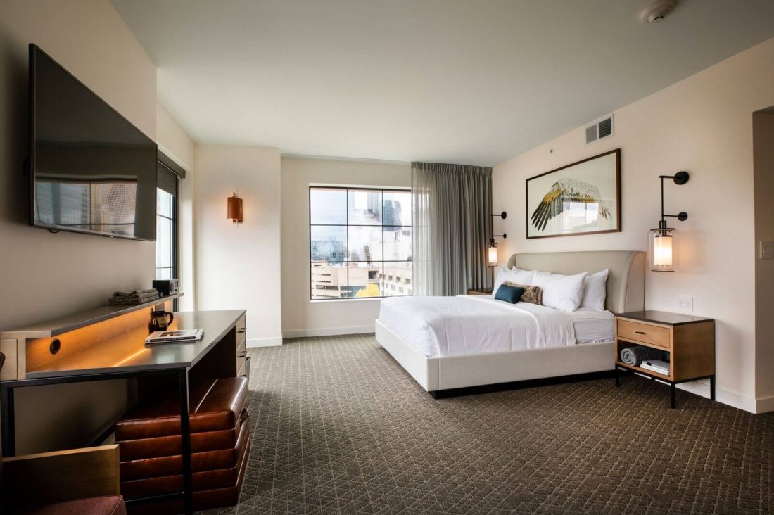 Modern hotel room with a large bed, desk, wall-mounted TV, nightstands, and a window with city view. The decor is minimalist with neutral tones and a framed artwork above the bed.