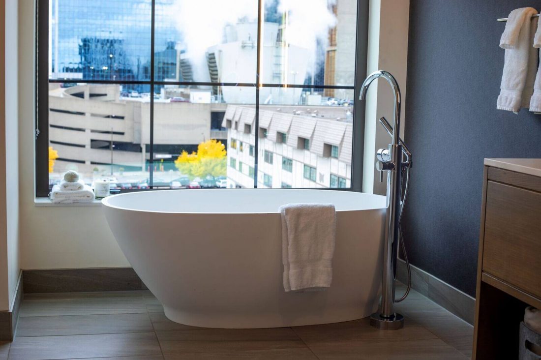 A modern white bathtub with a towel draped over the side sits by a large window overlooking city buildings.