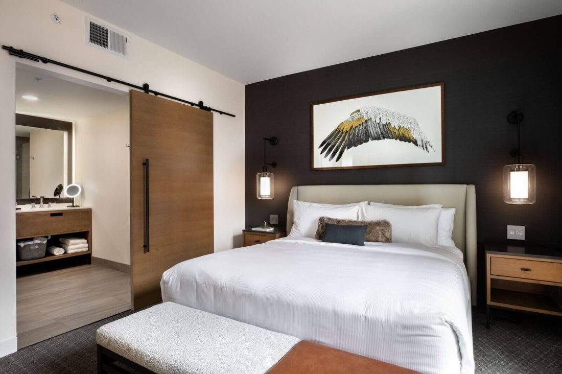 Modern hotel room with a neatly made bed, two bedside tables, a bench, wall lamps, and a large artwork of a bird above the headboard; bathroom visible through open sliding door.