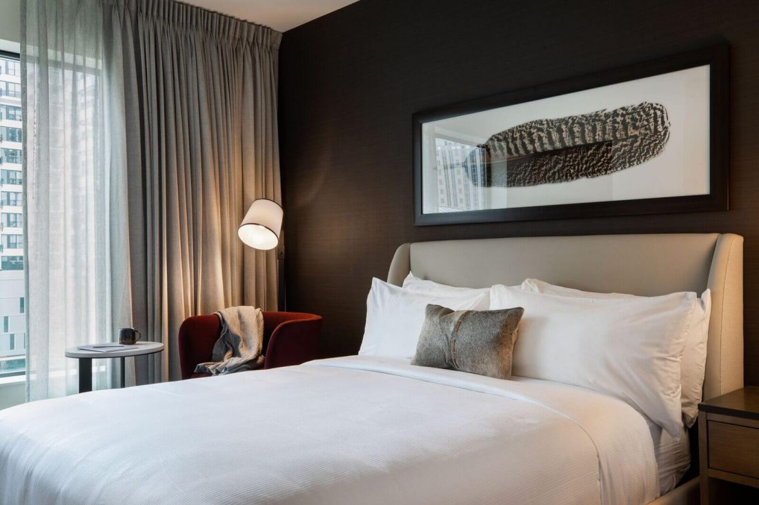 A modern Deluxe QUEEN hotel room with a neatly made bed, a large feather artwork above the headboard, a red chair by the window, and sheer curtains letting in natural light.