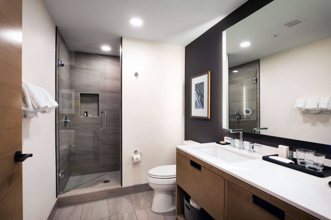 Modern bathroom in the Deluxe Queen features a glass-enclosed shower, toilet, large mirror, wooden vanity with double sinks, and towels neatly arranged on racks.