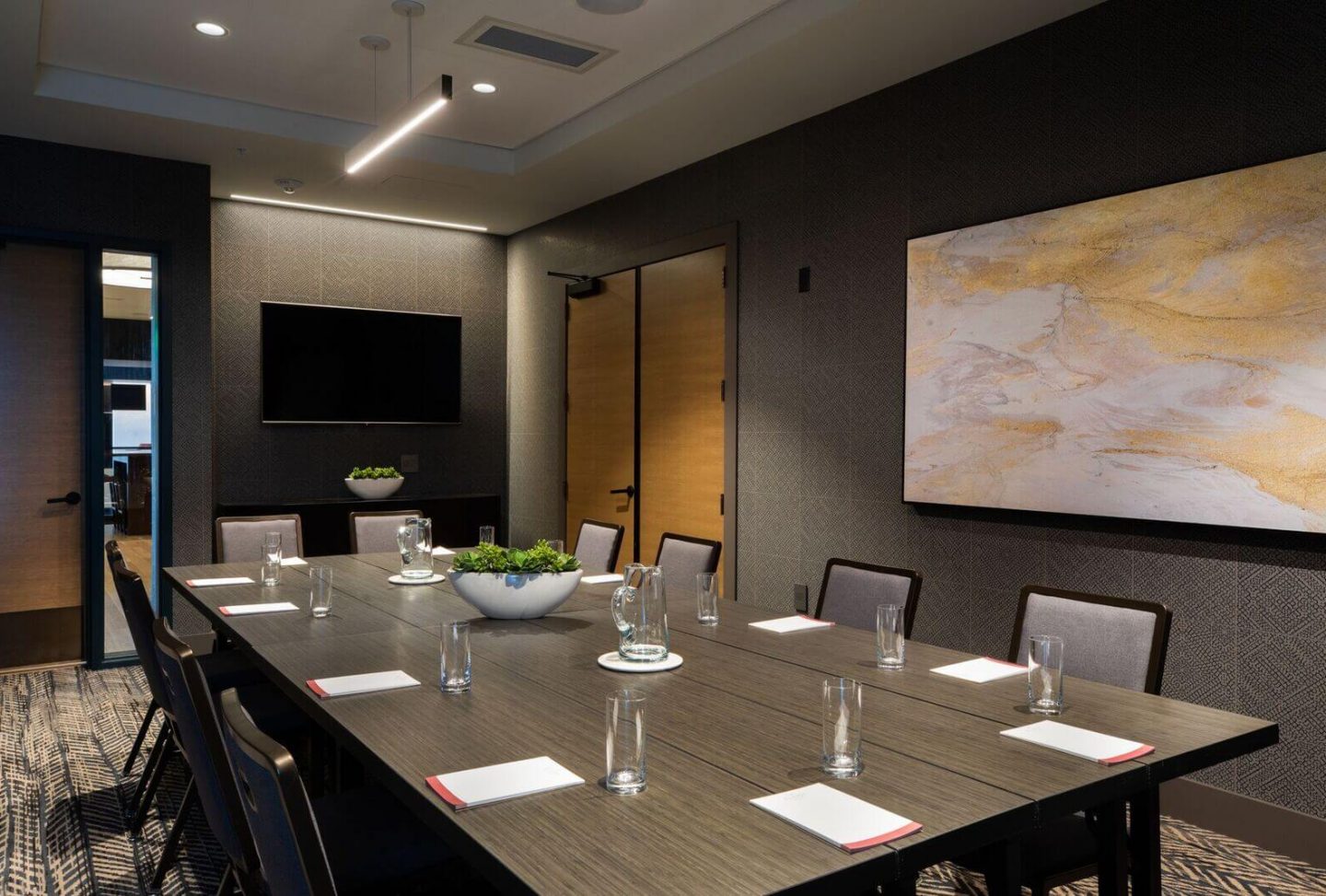 A modern conference room with a long table, eight chairs, notepads, glasses, a bowl of greenery, a wall-mounted TV, and abstract art on the wall.