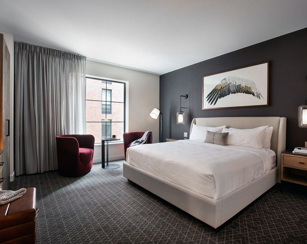 Modern hotel room with a large bed, dark accent wall, framed wing artwork, two red chairs by a window, and neutral-toned decor.