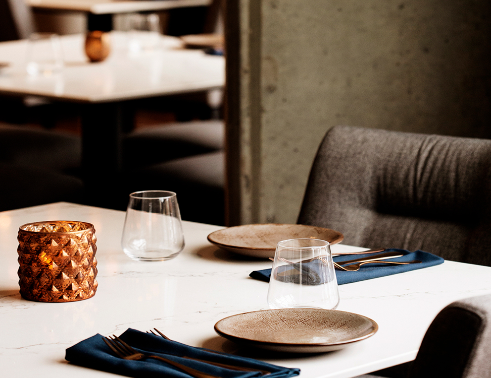 A restaurant table set for two with brown plates, blue napkins, gold utensils, clear glasses, and a decorative candle on a white marble surface.