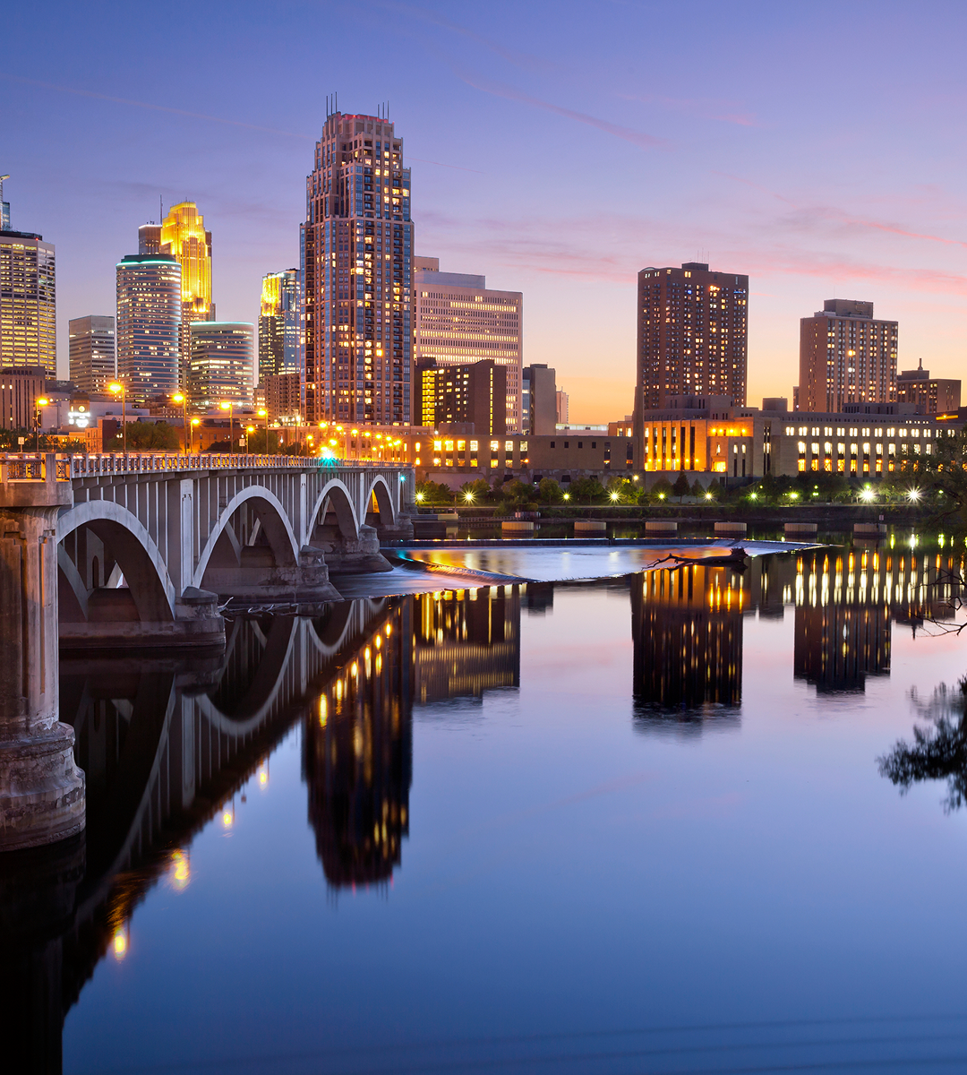 A city skyline at sunset with modern buildings, a bridge, and their reflections on a calm river—perfect for exploring new sights and discovering exciting things to do along the waterfront.
