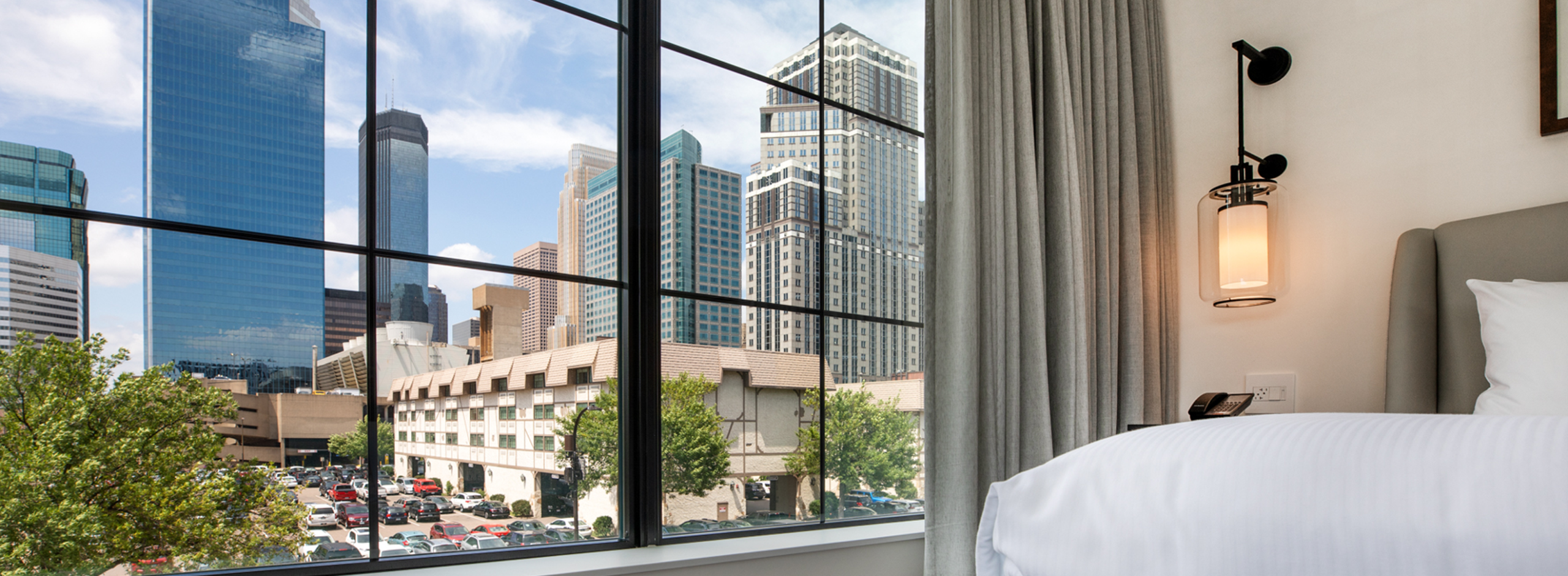 A city skyline with tall buildings is visible through a large window from a modern hotel room with white bedding and a wall lamp.