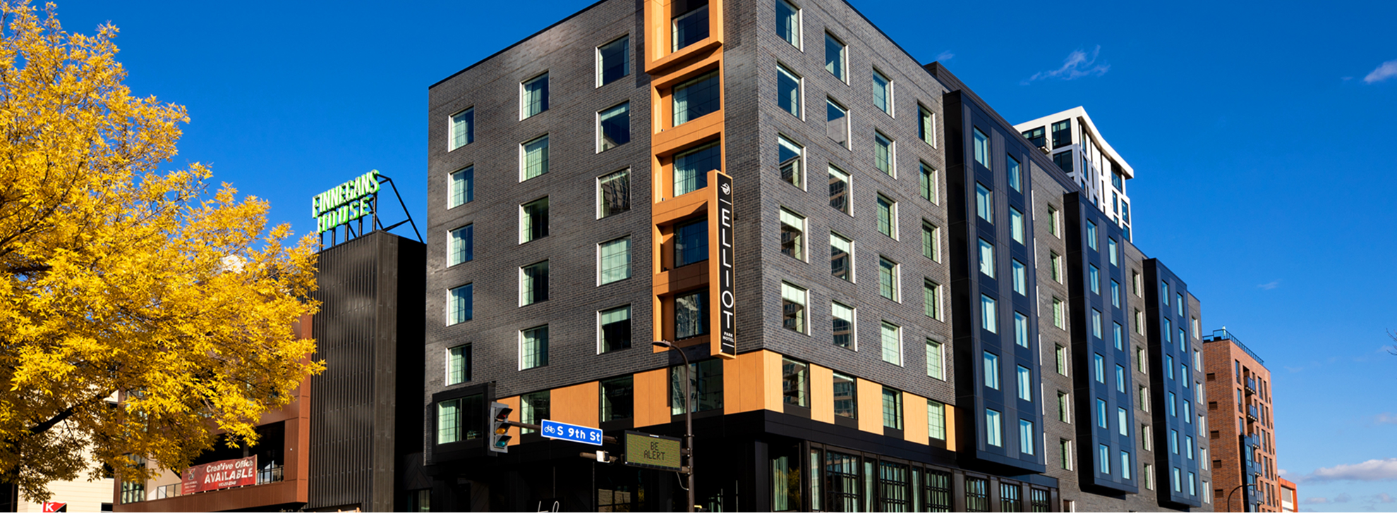 A modern mid-rise hotel building stands on a city street corner, its black and tan exterior and large windows highlighting the prime location beneath a clear blue sky.