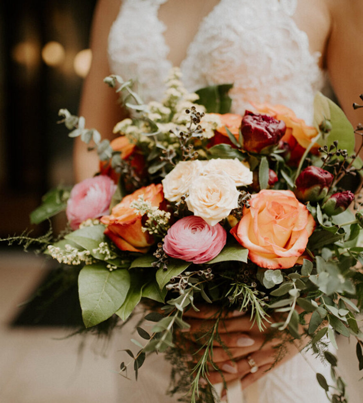 A person in a white lace dress holds a bouquet of roses and various flowers in shades of orange, pink, and cream, with green foliage.