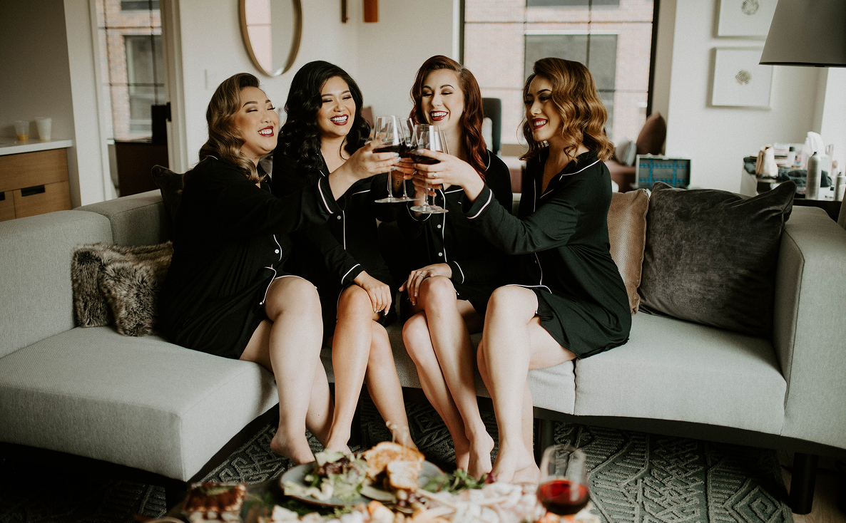 Four women in matching black pajamas sit on a sofa, smiling and clinking wine glasses, with a tray of food and drinks on the table in front of them.
