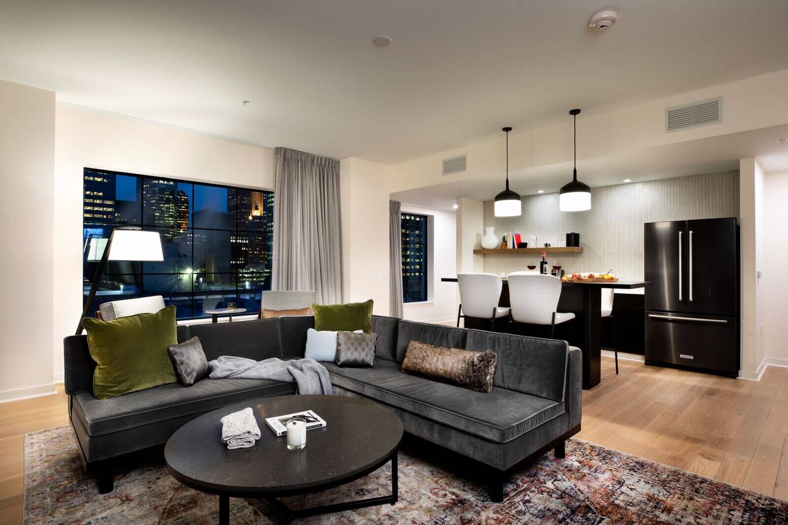 Modern open-concept living room and kitchen with a dark sectional sofa, round coffee table, and city view through large window at night.