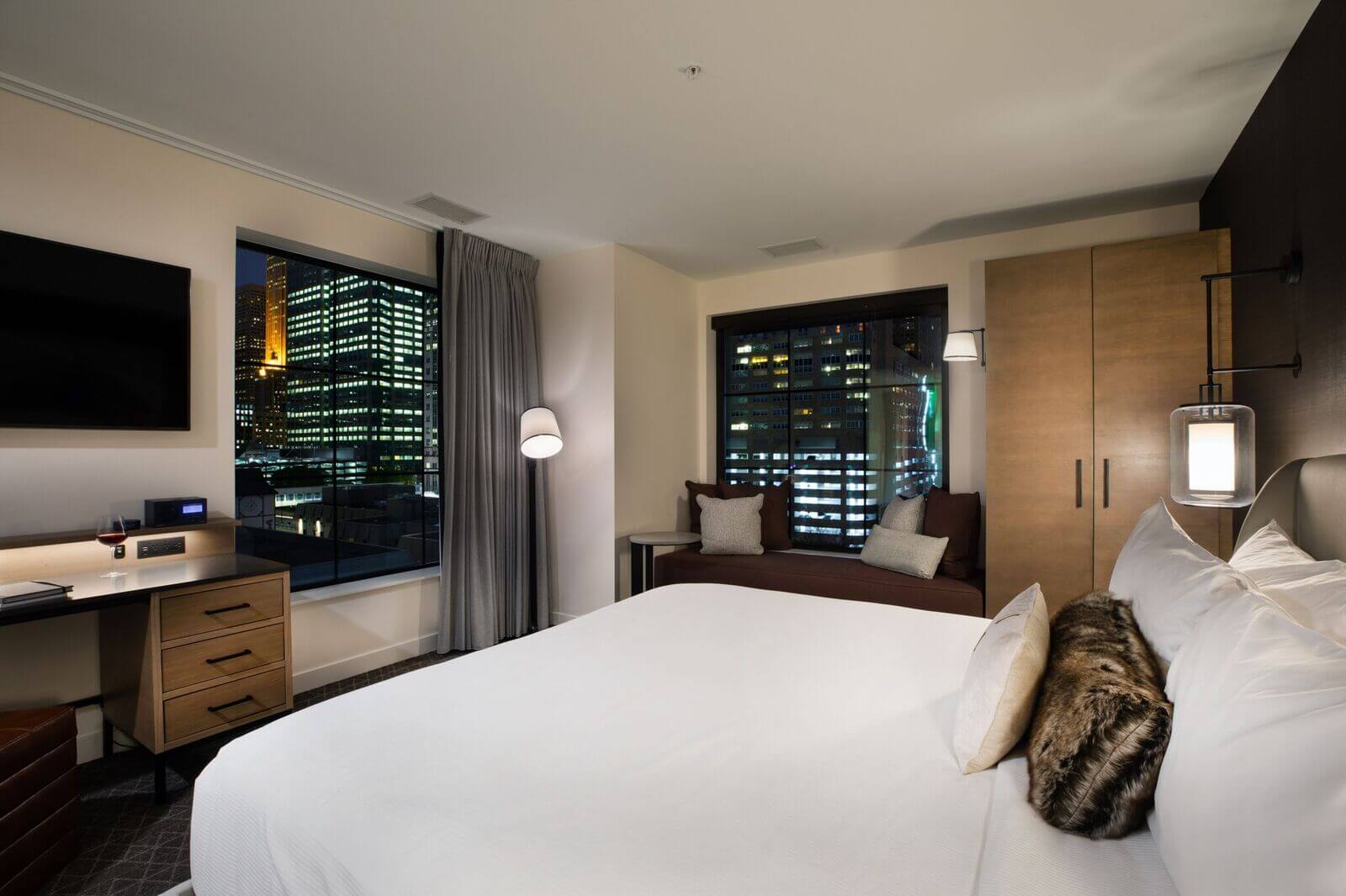 Modern hotel room with a large bed, desk, sofa, and cityscape views through two windows at night; room features neutral tones and contemporary furnishings.