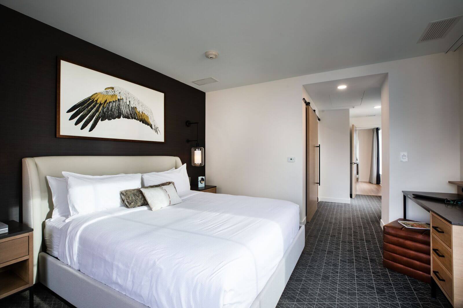 A modern hotel room with a neatly made bed, a large bird wing artwork above the headboard, bedside tables, and a view into a hallway with natural light.