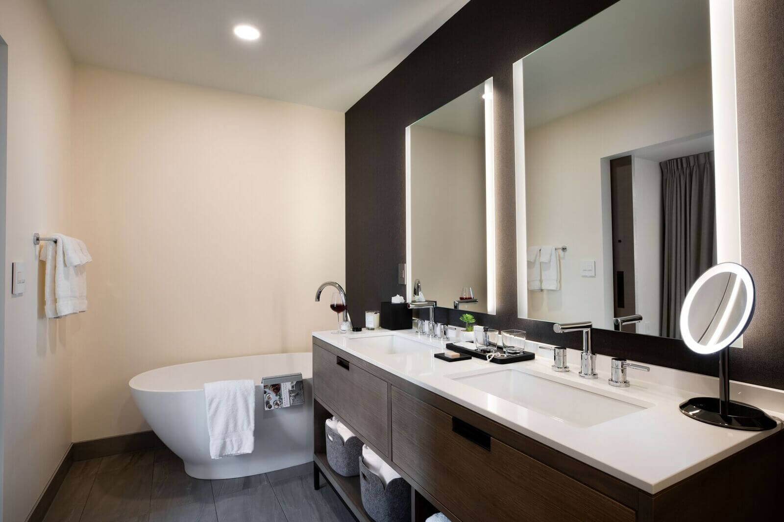 Modern bathroom with a freestanding bathtub, double-sink vanity, illuminated mirrors, countertop toiletries, towel racks, and neutral color palette.