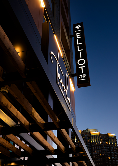 Exterior view of Elliot Park Hotel at dusk, featuring illuminated signage and wooden architectural elements with a city building in the background.