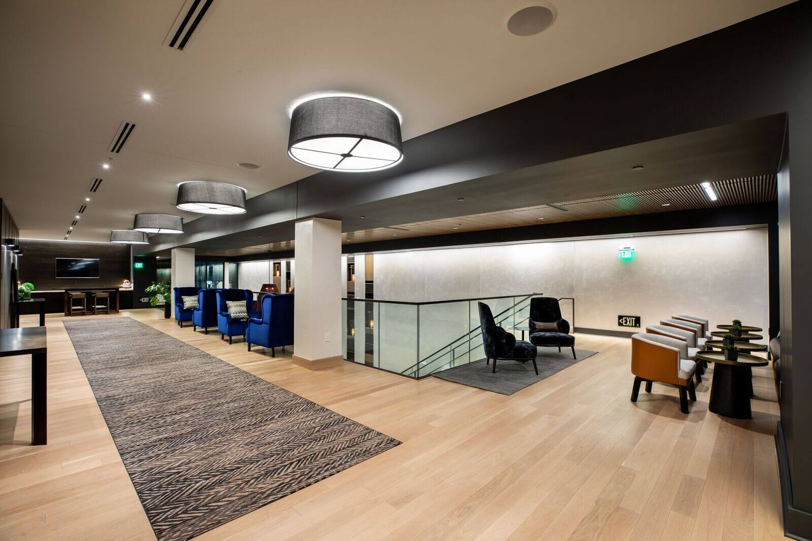 Modern lounge pre-function space with wood flooring, armchairs, and overhead lighting, featuring a glass railing by the staircase and a carpeted walkway.