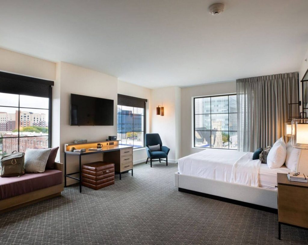 Modern hotel room with a large bed, desk, bench seating, armchair, TV, and large windows overlooking a cityscape.