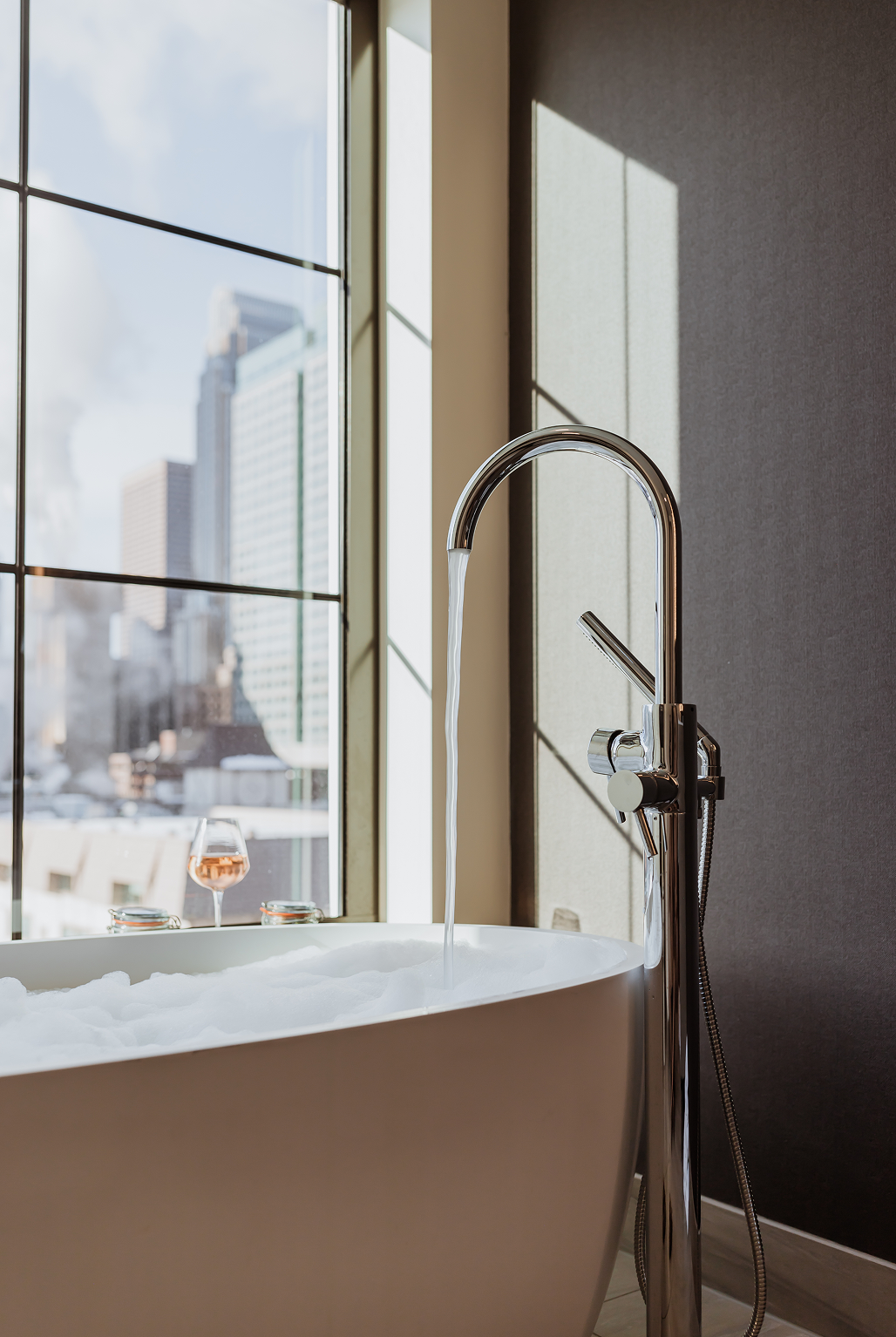 A modern bathtub with running water and bubbles sits by large windows overlooking a cityscape; a glass of wine and candles rest on the tubs edge.