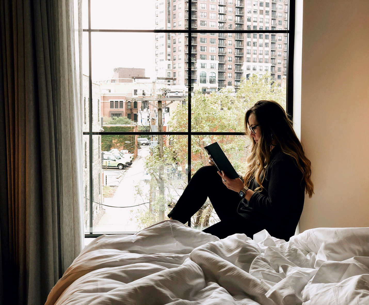 A person sits on a windowsill reading a book, with an urban cityscape visible outside. A bed with white blankets is in the foreground.