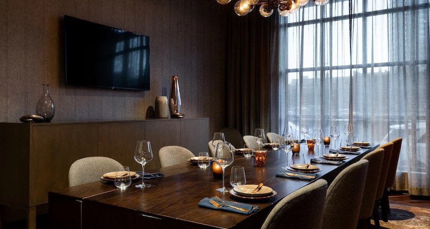 Experience private dining in this modern Tavola-inspired dining room, featuring a long wooden table for eight, upholstered chairs, pendant lighting, decorative vases, and large windows with sheer curtains.