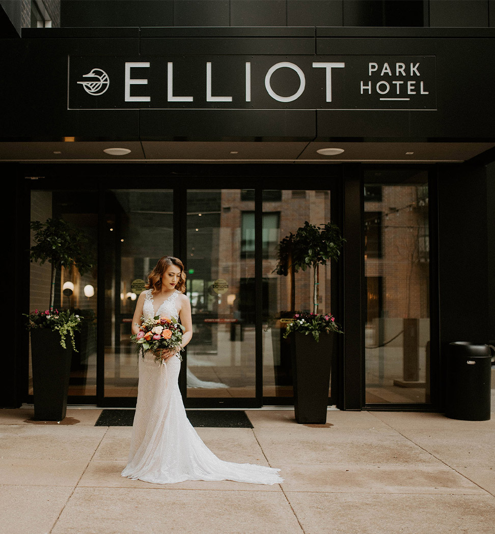 A bride in a white wedding dress stands holding a bouquet outside the entrance of Elliot Park Hotel.